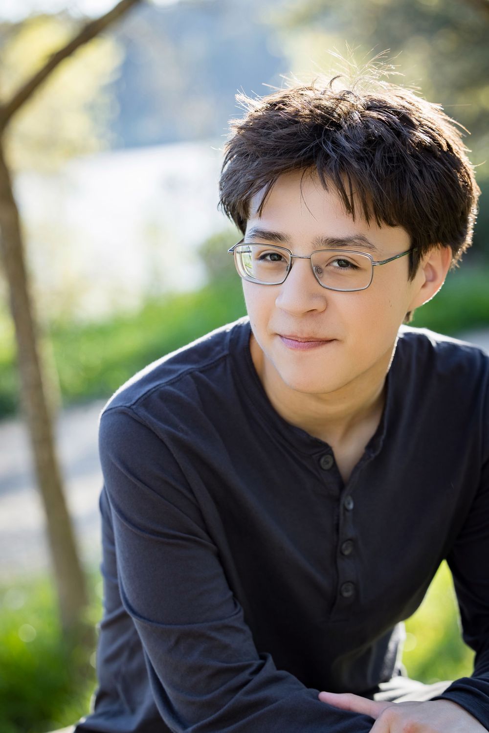 A person wearing glasses and a dark shirt smiles warmly while sitting outdoors in natural lighting.