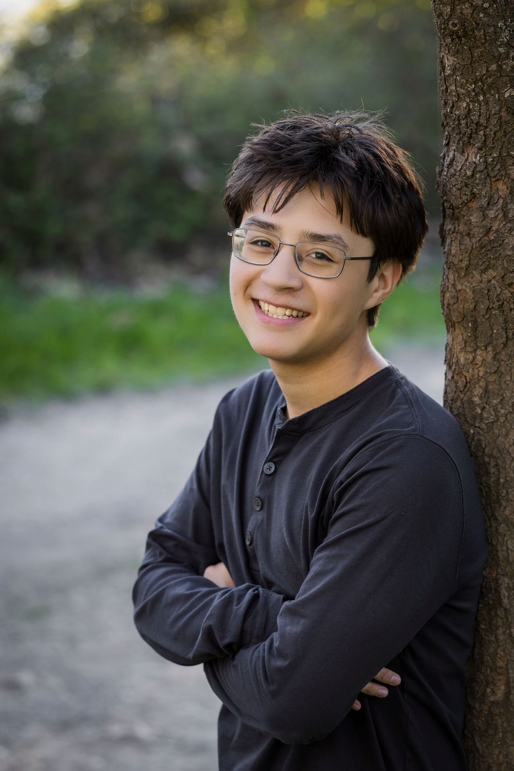 A person wearing a black sweater leans against a tree trunk outdoors while smiling warmly in a natural setting.
