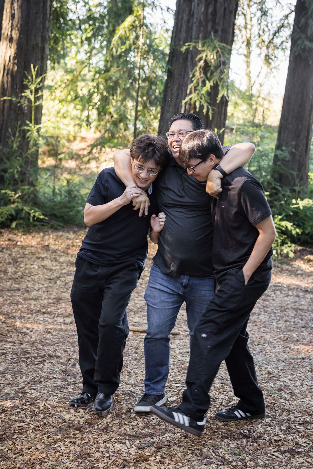 Three people wearing black clothing pose playfully together in a wooded outdoor setting.