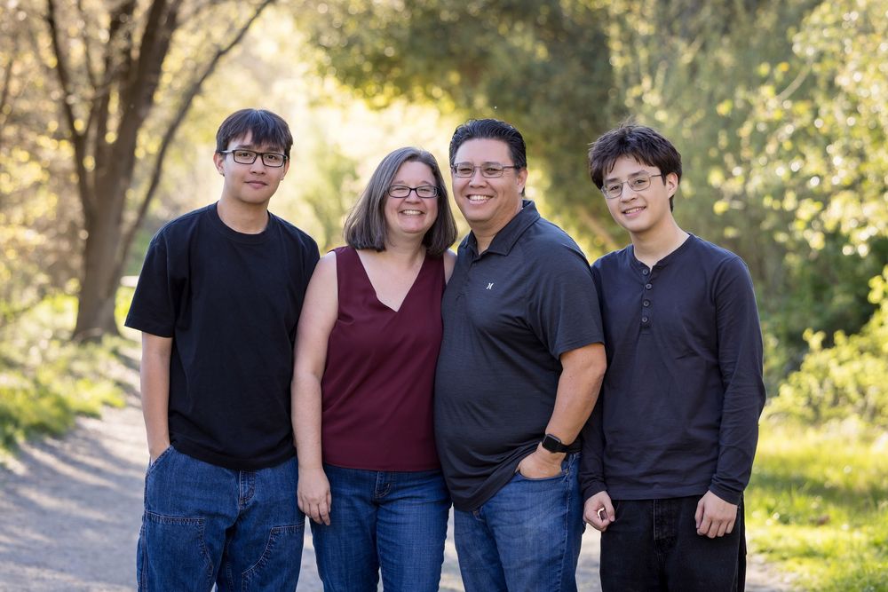 A group of people in casual attire pose together outdoors in front of sunlit trees and foliage.