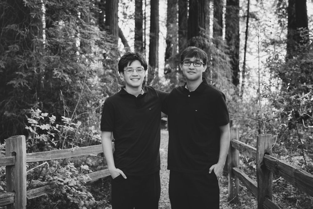 Two people in black shirts stand together on a wooden walkway in a peaceful forest setting.