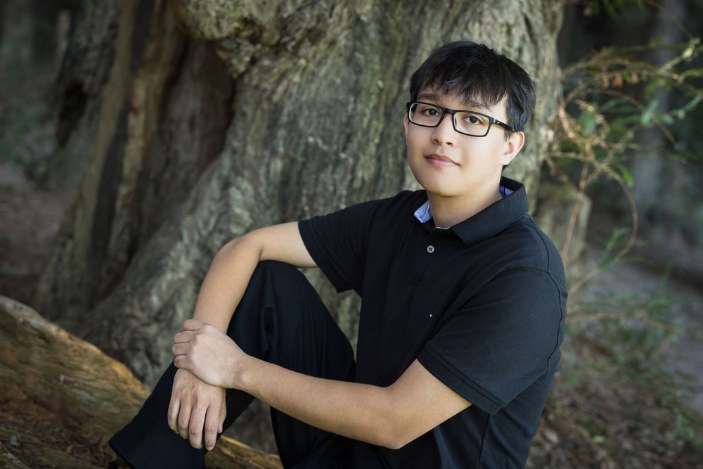 Person in black hoodie and glasses poses in outdoor wooded setting with natural lighting.