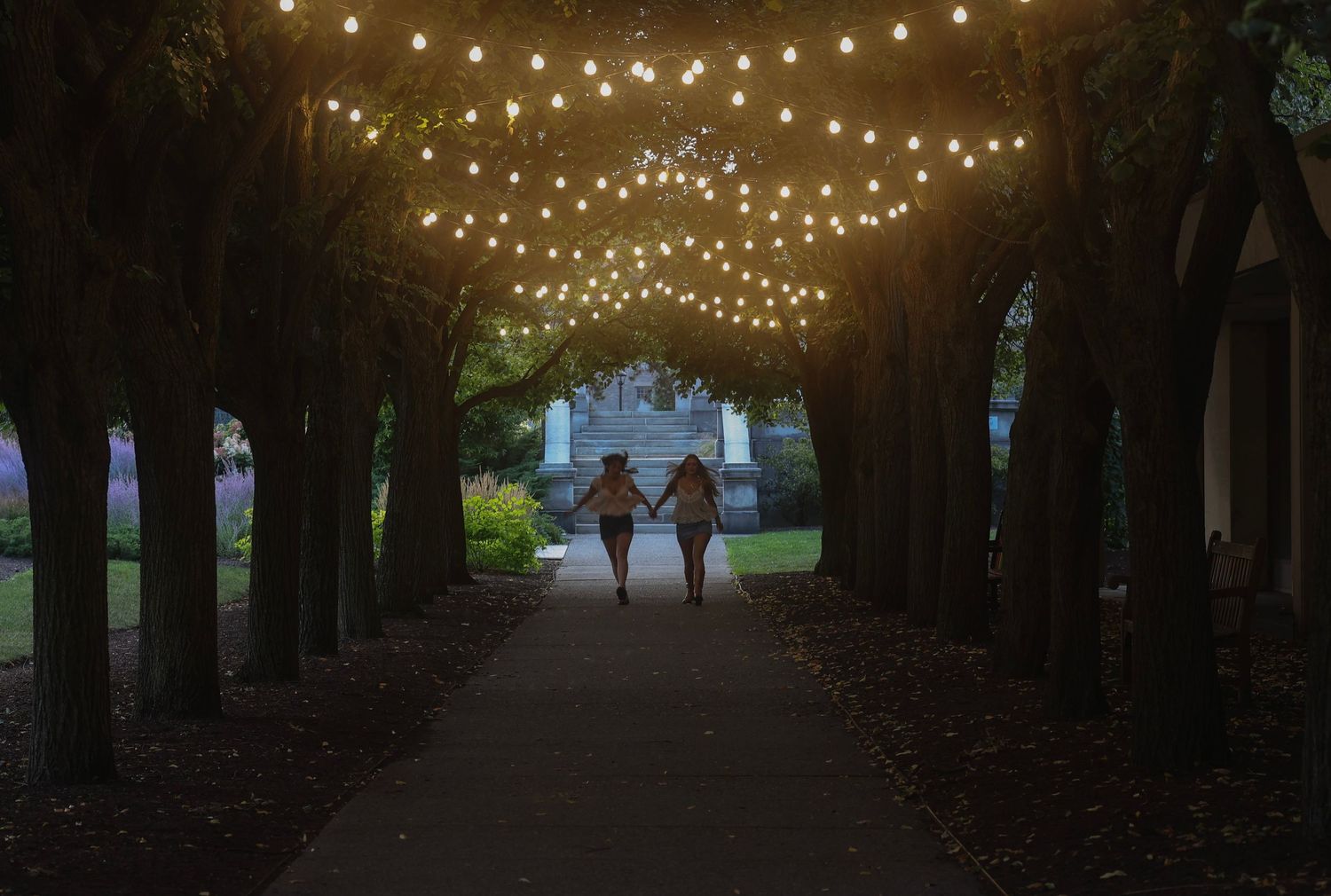 Two women running under a walkway with string lights overhead