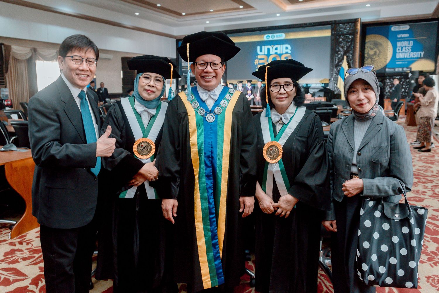 A group of graduates in blue academic regalia and guests pose together for celebratory photos at a graduation ceremony.