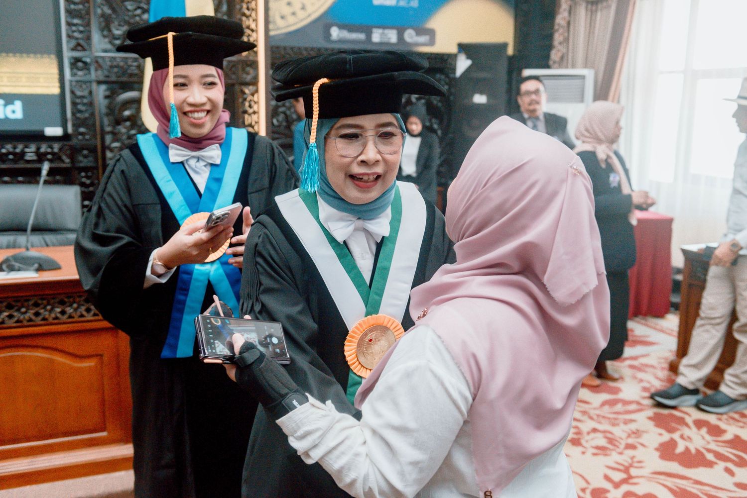 Graduates in blue and white academic regalia celebrate during a university commencement ceremony.
