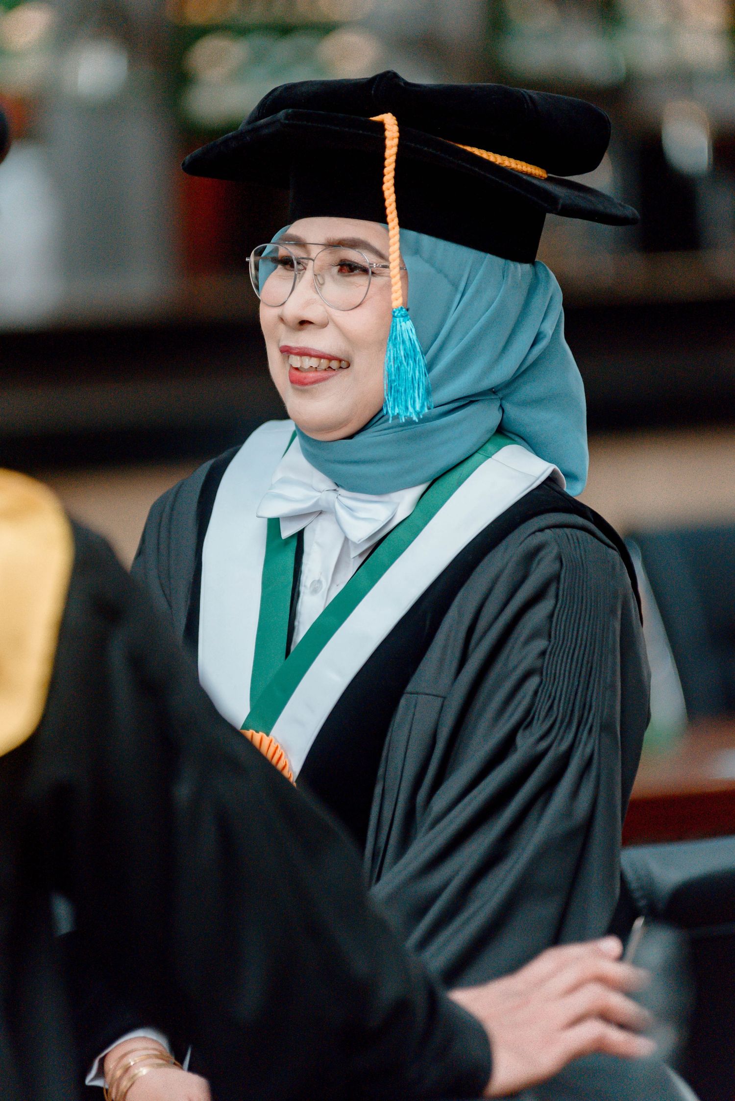 Graduate in black cap and gown with green stole smiles during commencement ceremony.