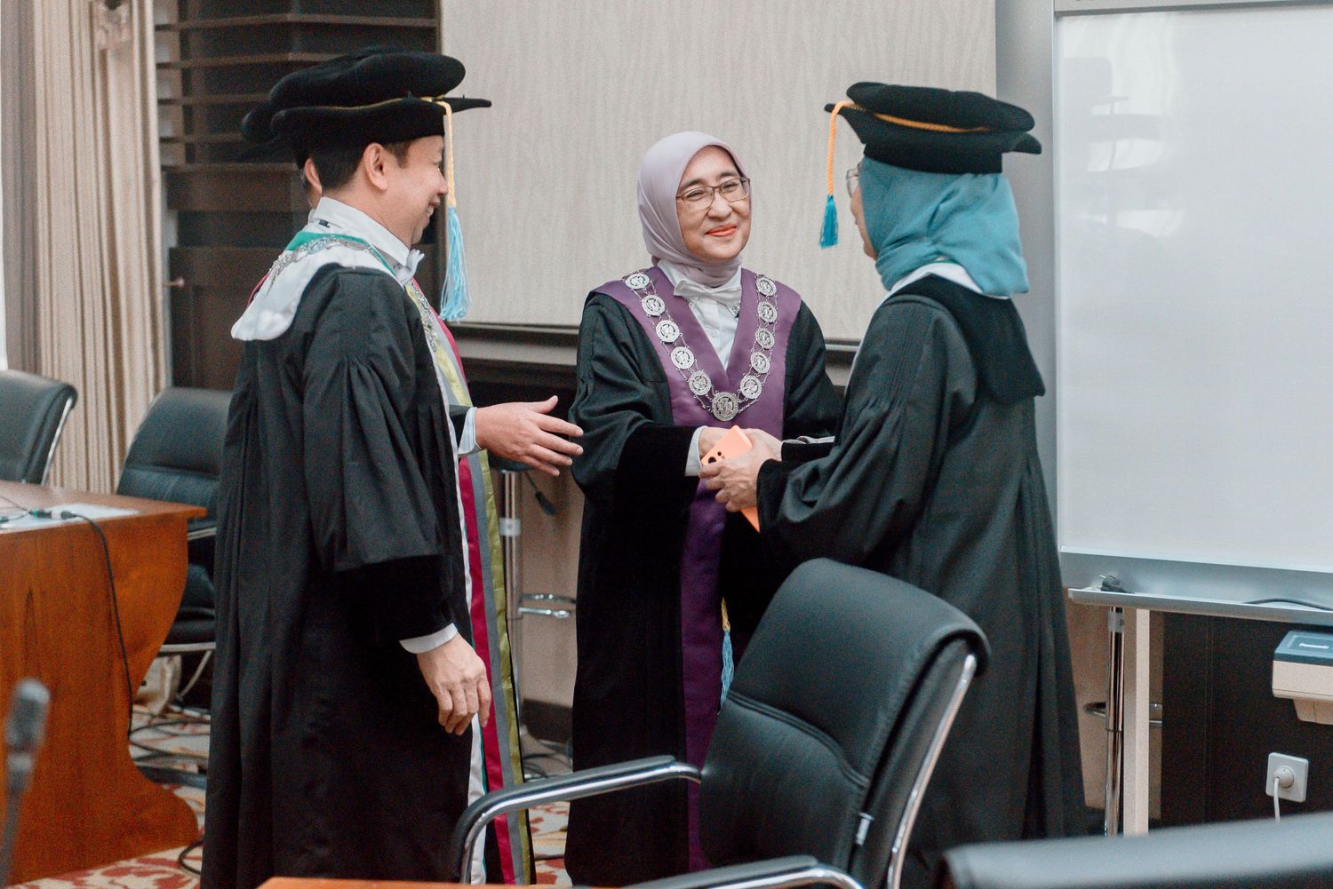 Three people in academic graduation regalia interact during a ceremony in an indoor setting.