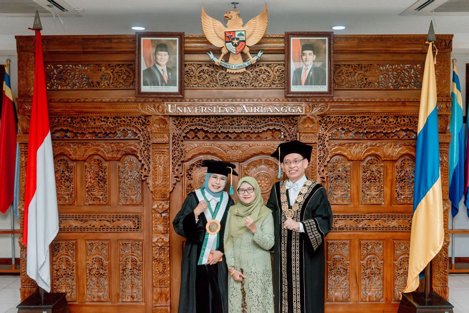 Graduation ceremony photo with Indonesian national emblem and ornate wooden carved wall panel backdrop.