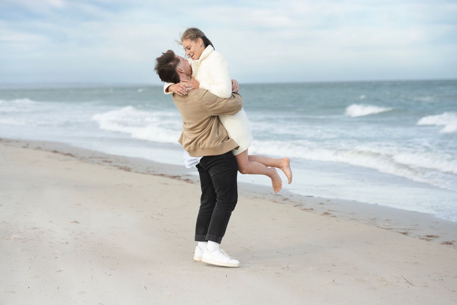 A man lifts a woman joyfully on a sandy beach with ocean waves in the background.