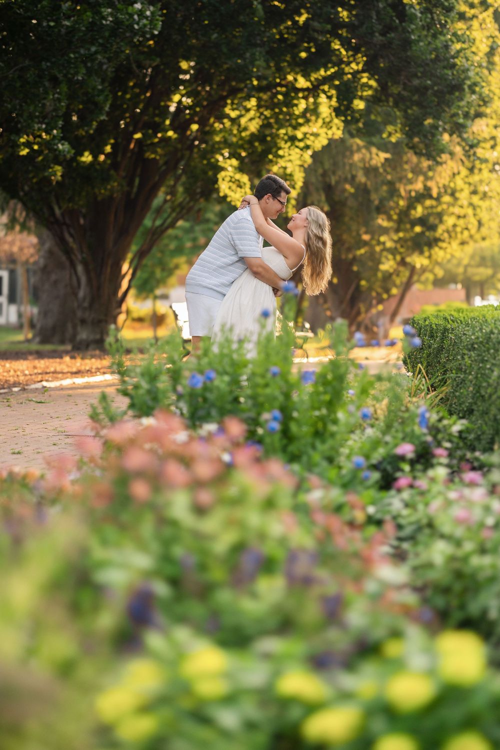 A couple shares a romantic kiss in a lush garden setting with blooming flowers and golden sunlight filtering through trees. German Village