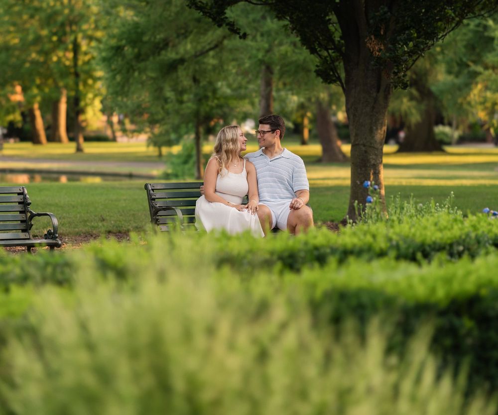 A couple sits together on a park bench surrounded by lush greenery and trees during golden hour. German Village Engagement session