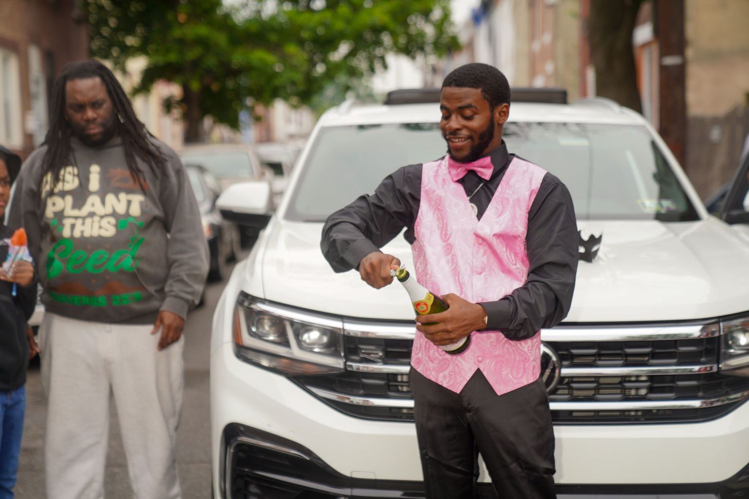 A person in a pink vest checks their watch while standing in front of a white SUV on a city street.