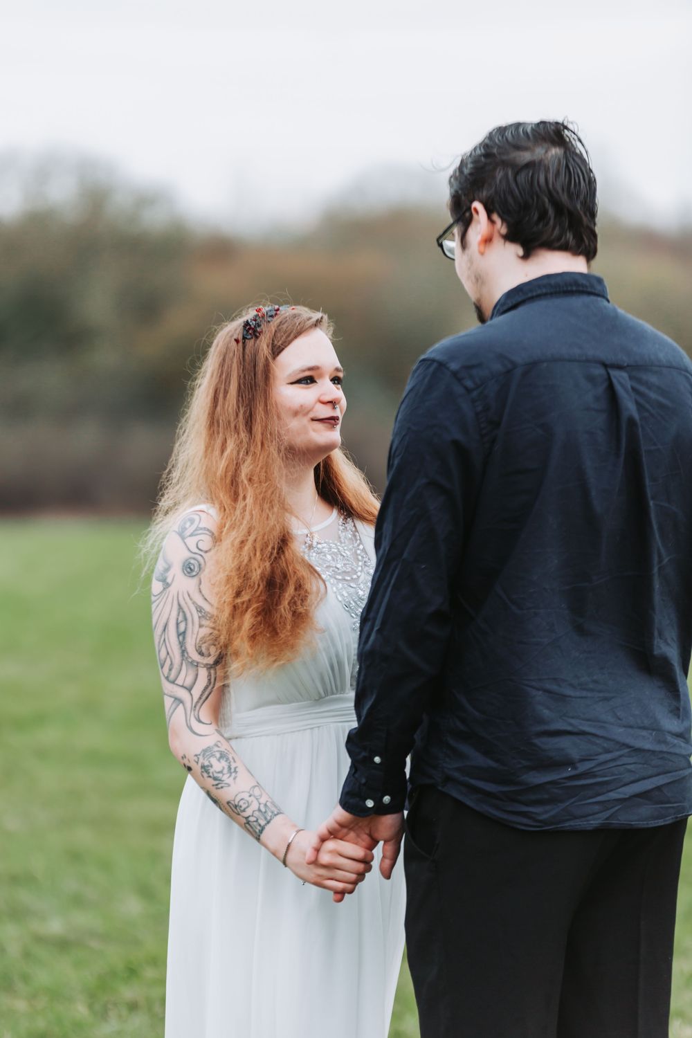 A couple in wedding attire walks together in a green field on an overcast day.