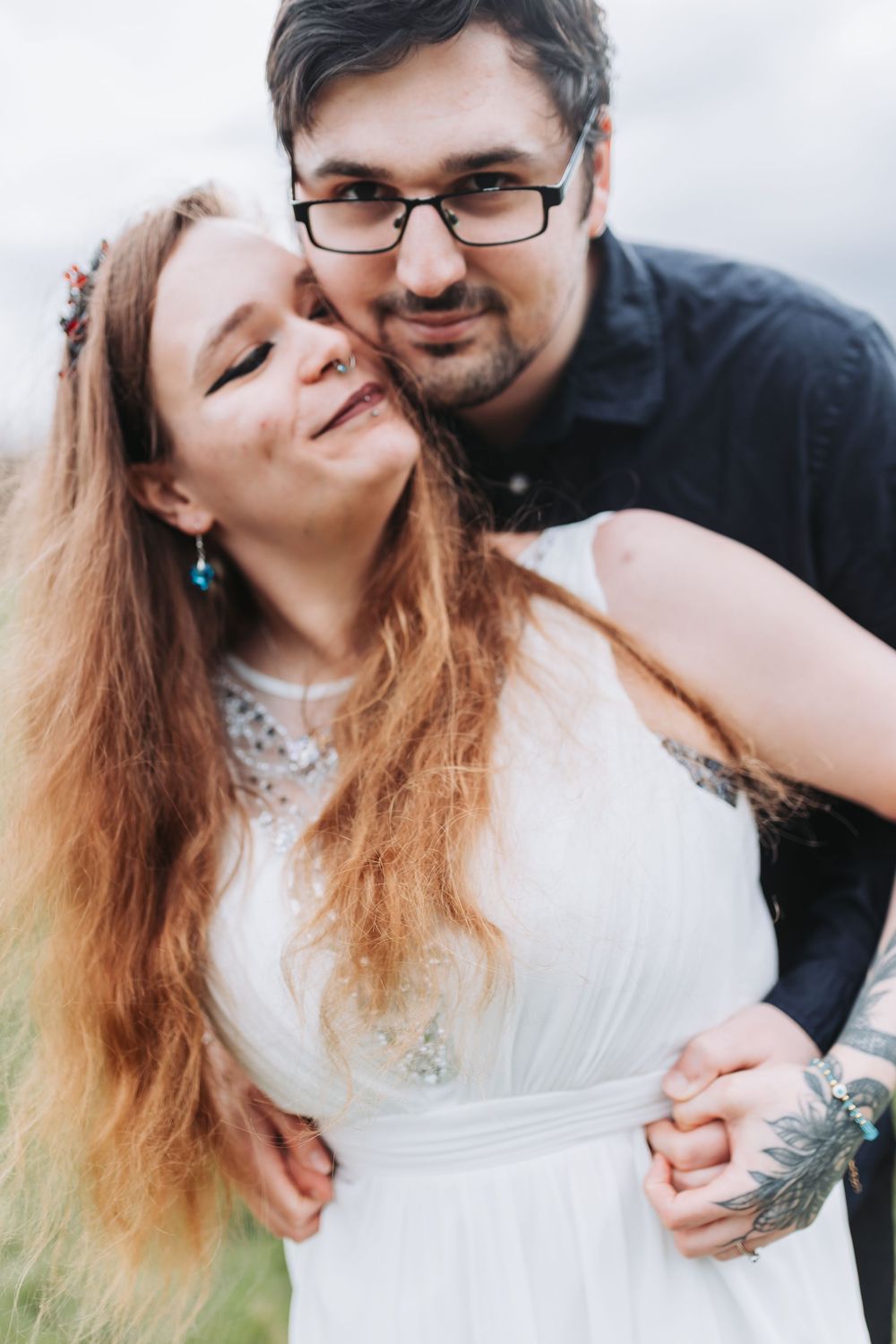 A happy couple embraces outdoors, with the bride wearing a white dress and the groom in black with glasses.