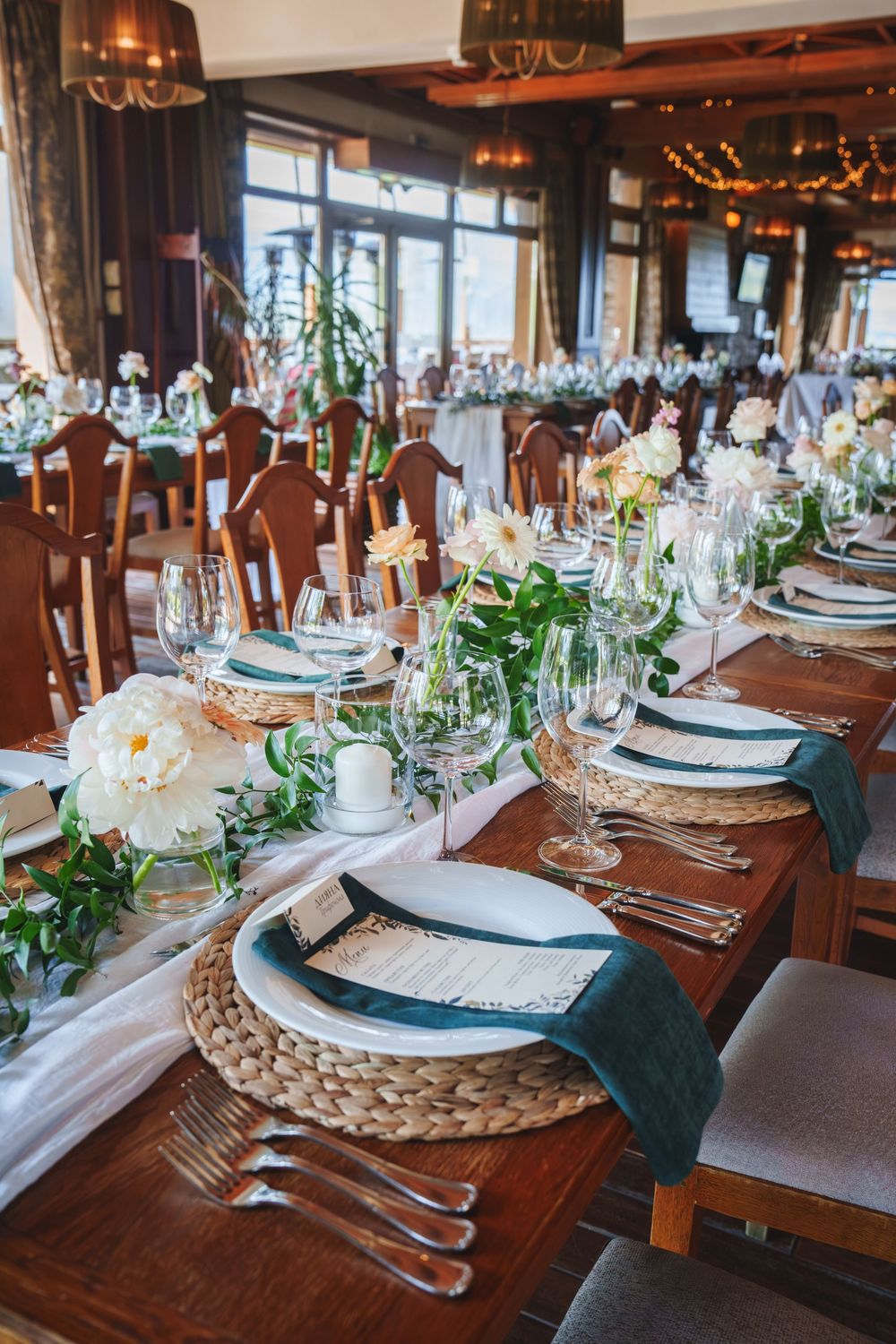 Elegant table settings with woven chargers, blue napkins, and greenery on a rustic wooden dining table at an upscale event.