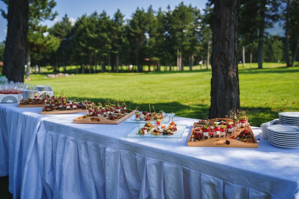Outdoor buffet table with white linens and serving dishes set up near trees and grass on a sunny day.