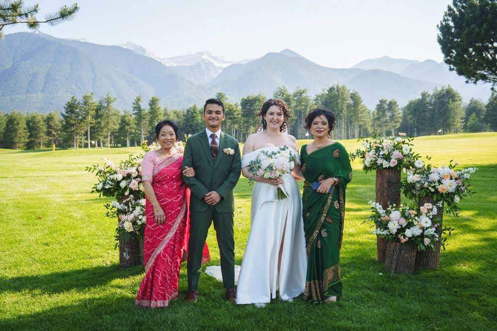 Wedding party poses on a grassy lawn with snow-capped mountains and evergreen trees in the background.