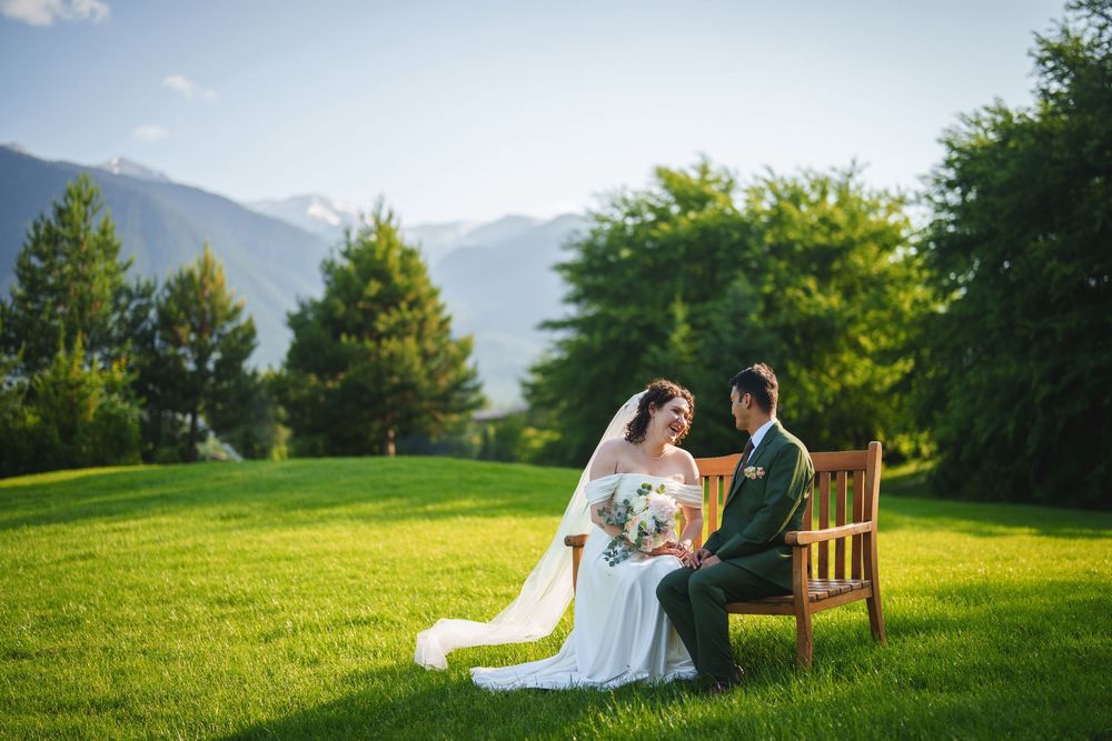 A romantic wedding scene on a wooden bench surrounded by lush green mountains and trees on a sunny day.