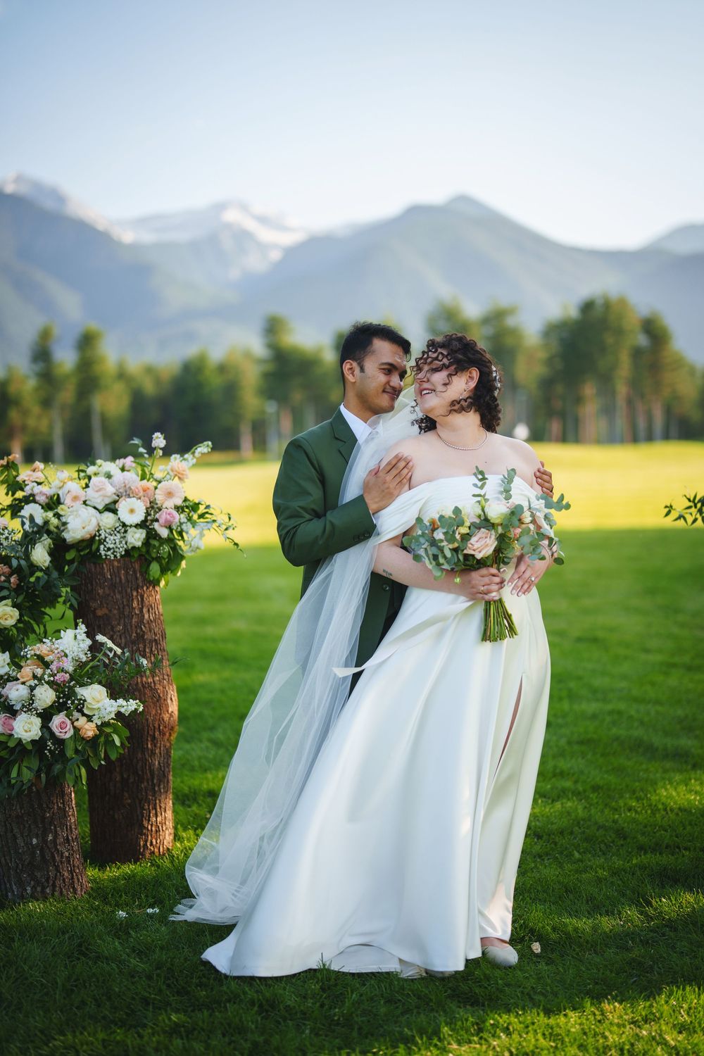 A wedding couple shares romantic moments in a scenic mountain meadow with white floral bouquets and green grass.