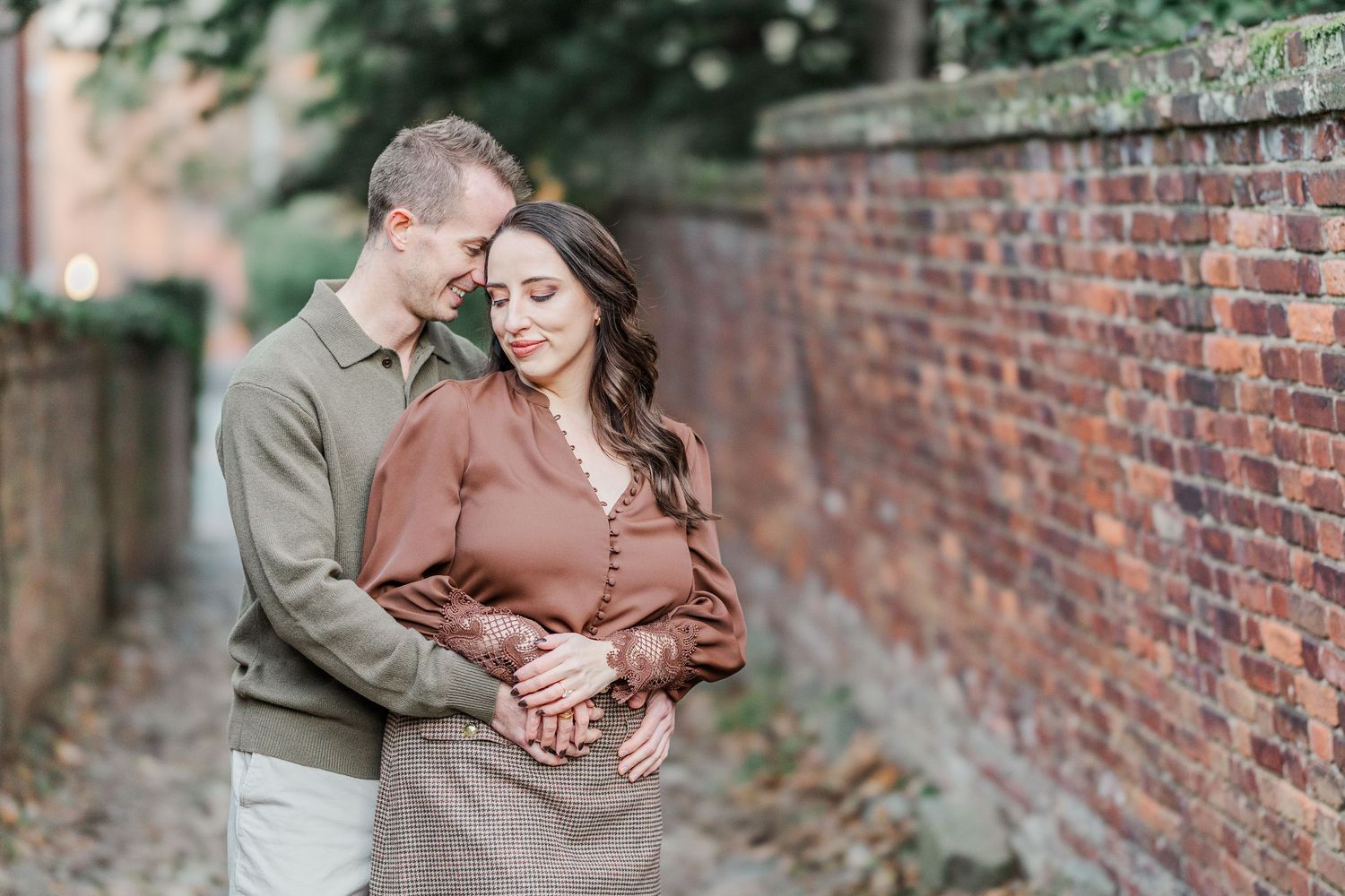A romantic couple embraces by a rustic brick wall during an outdoor autumn photoshoot.