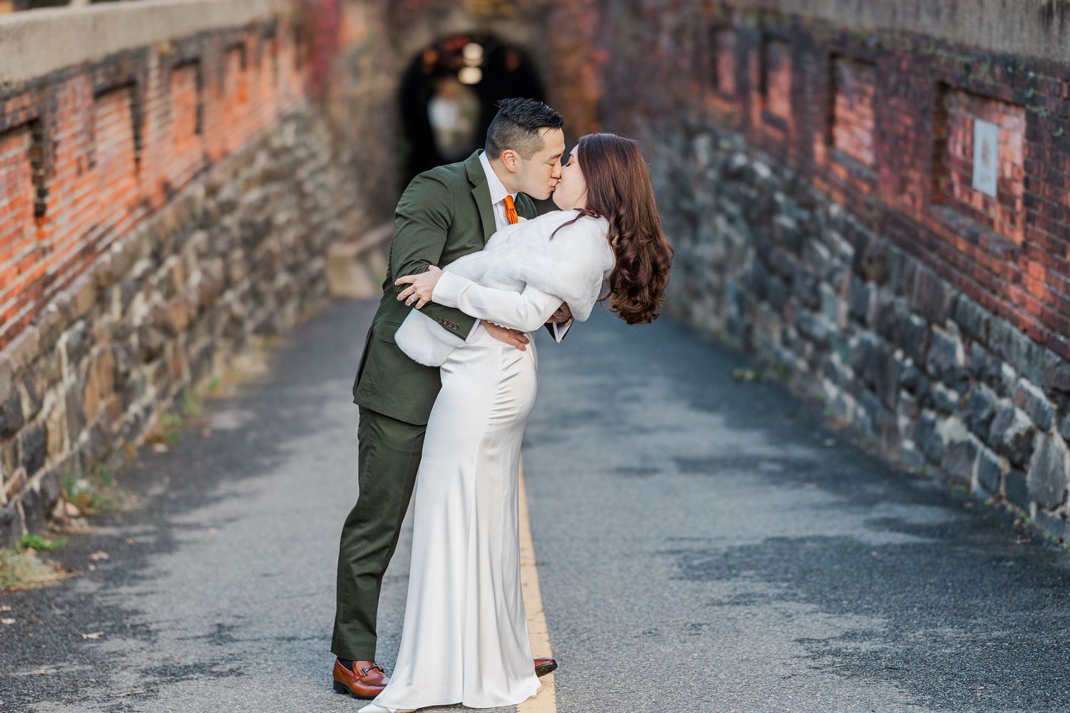 A couple shares a romantic dip kiss in front of a rustic brick wall.
