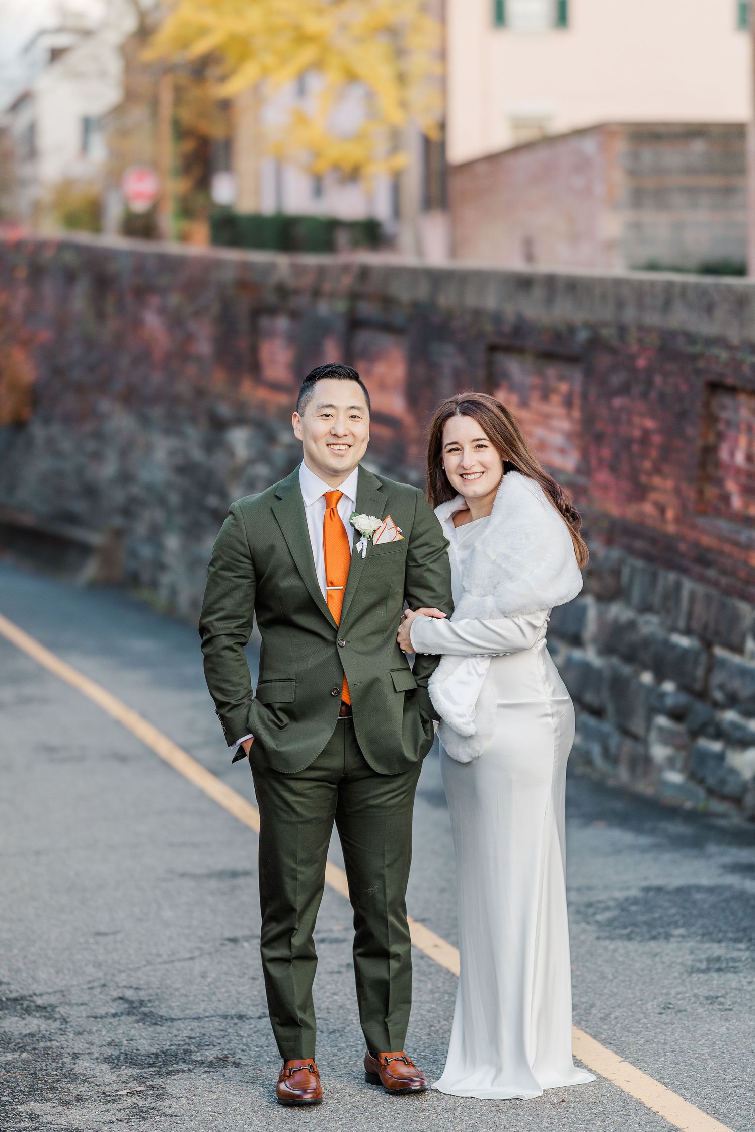 A couple in wedding attire stands on a brick walkway during autumn with warm lighting and fall foliage in the background.