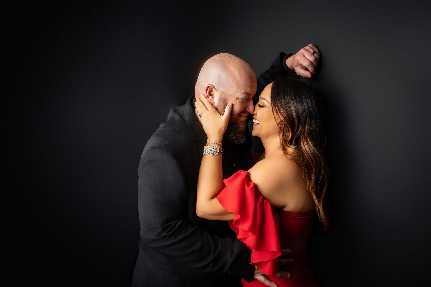 A romantic couple shares an intimate moment against a dark background as she wears a vibrant red dress.