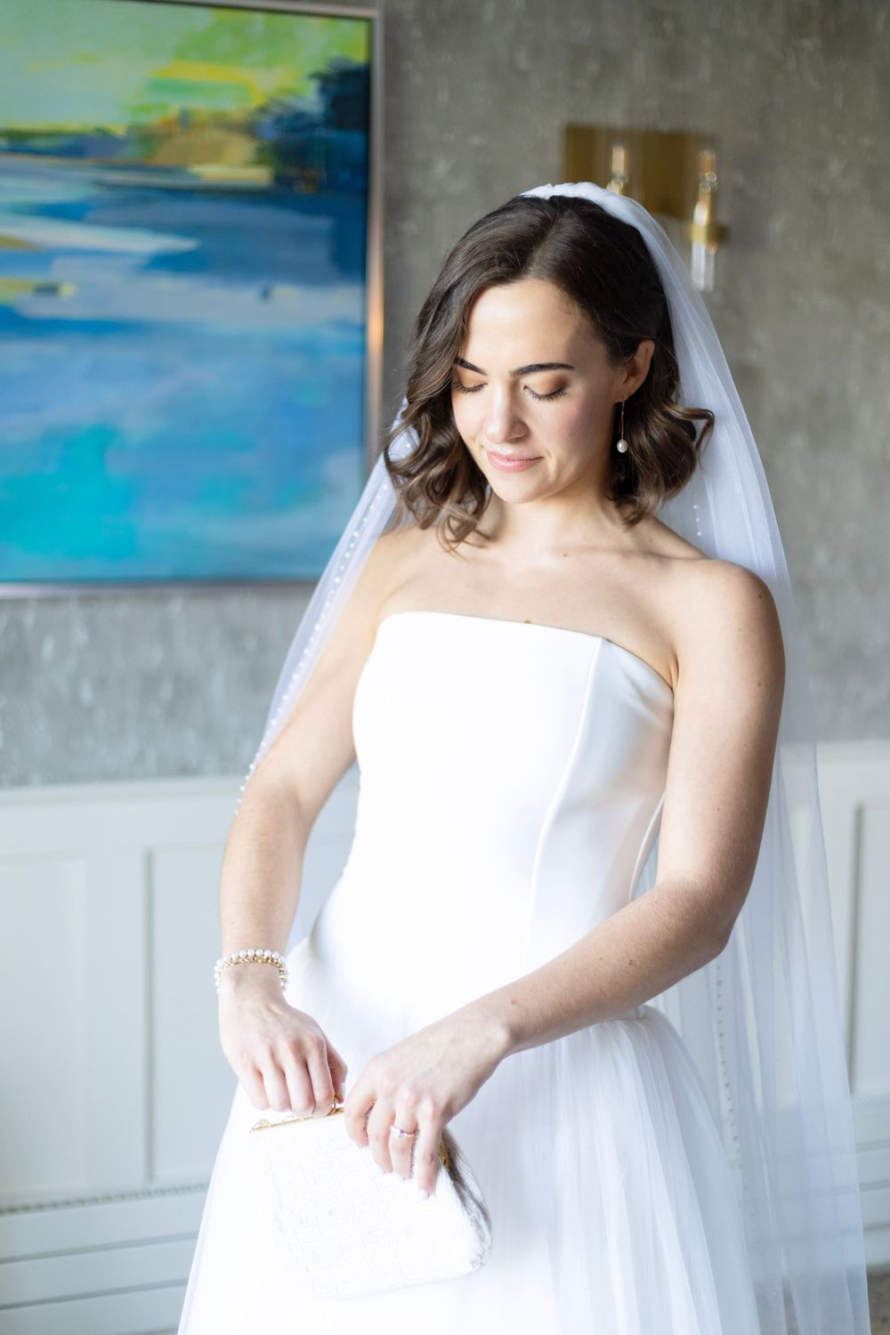 A bride in a white wedding gown and veil looks down thoughtfully in an elegant room with coastal artwork.
