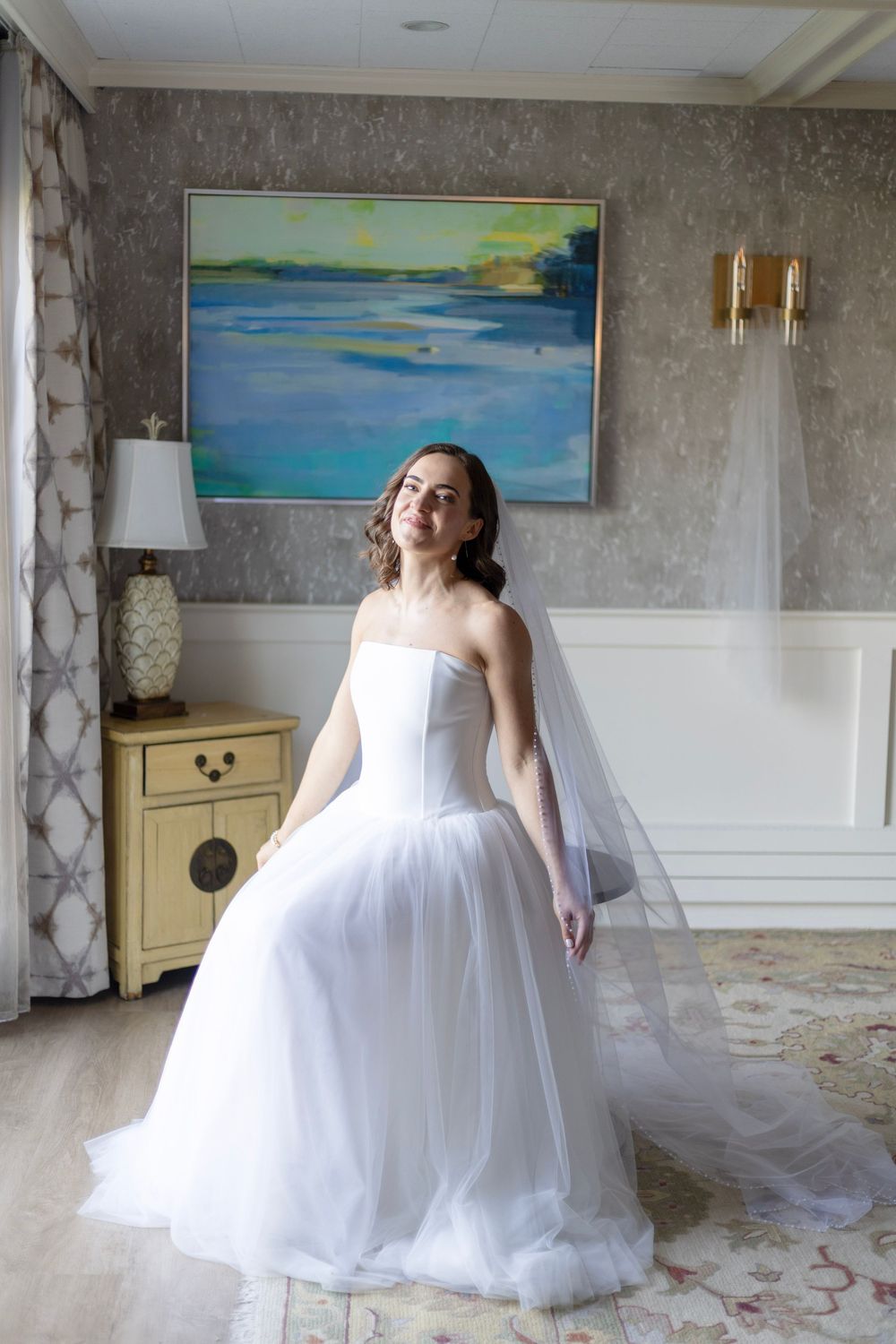 A flowing white wedding dress with tulle skirt is displayed in a room with coastal artwork on concrete walls.