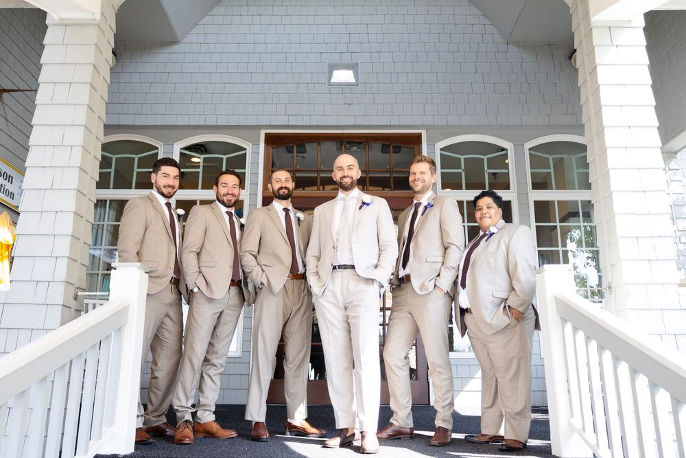 Wedding party in light beige suits poses on white porch steps for formal group photo.