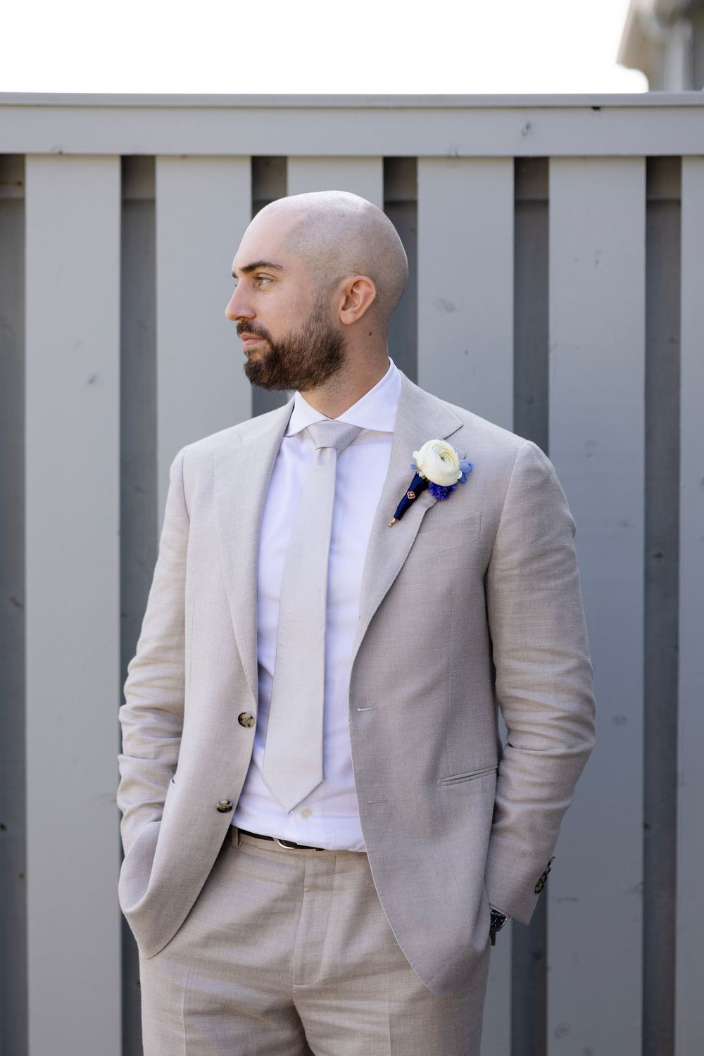 A groom in a light beige suit and white shirt with boutonniere poses against a gray wall during a wedding.