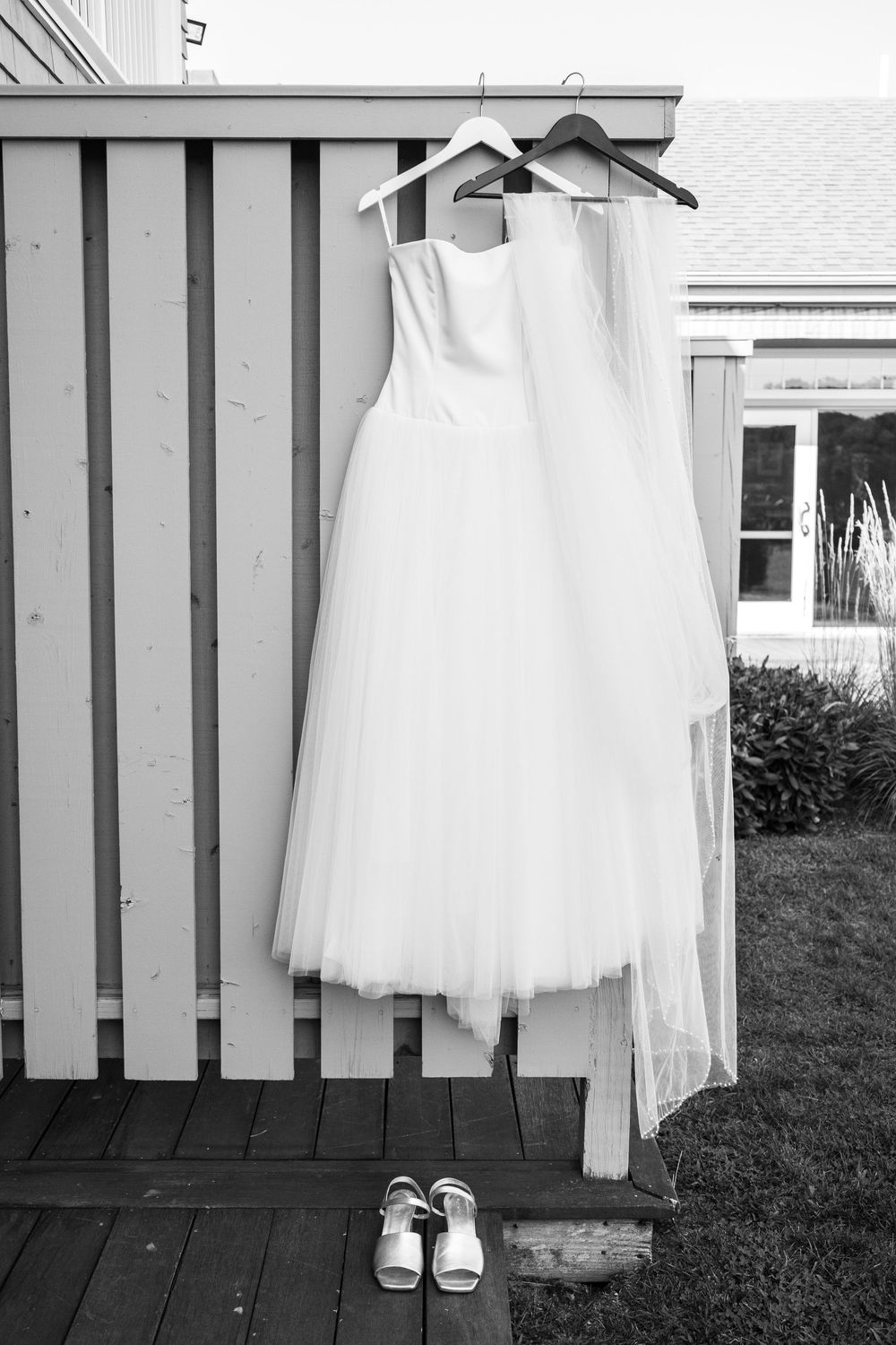 A white wedding dress and veil hang on a wooden deck with white shoes placed beneath it in black and white.