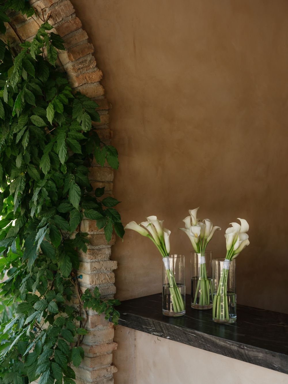 Elegant white calla lilies arranged in glass vases beside a stone archway with lush green foliage.