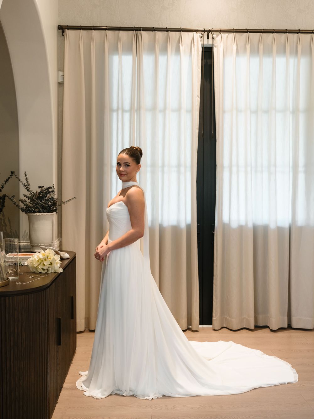 An elegant white wedding dress with a flowing train photographed in a bright room with sheer curtains.