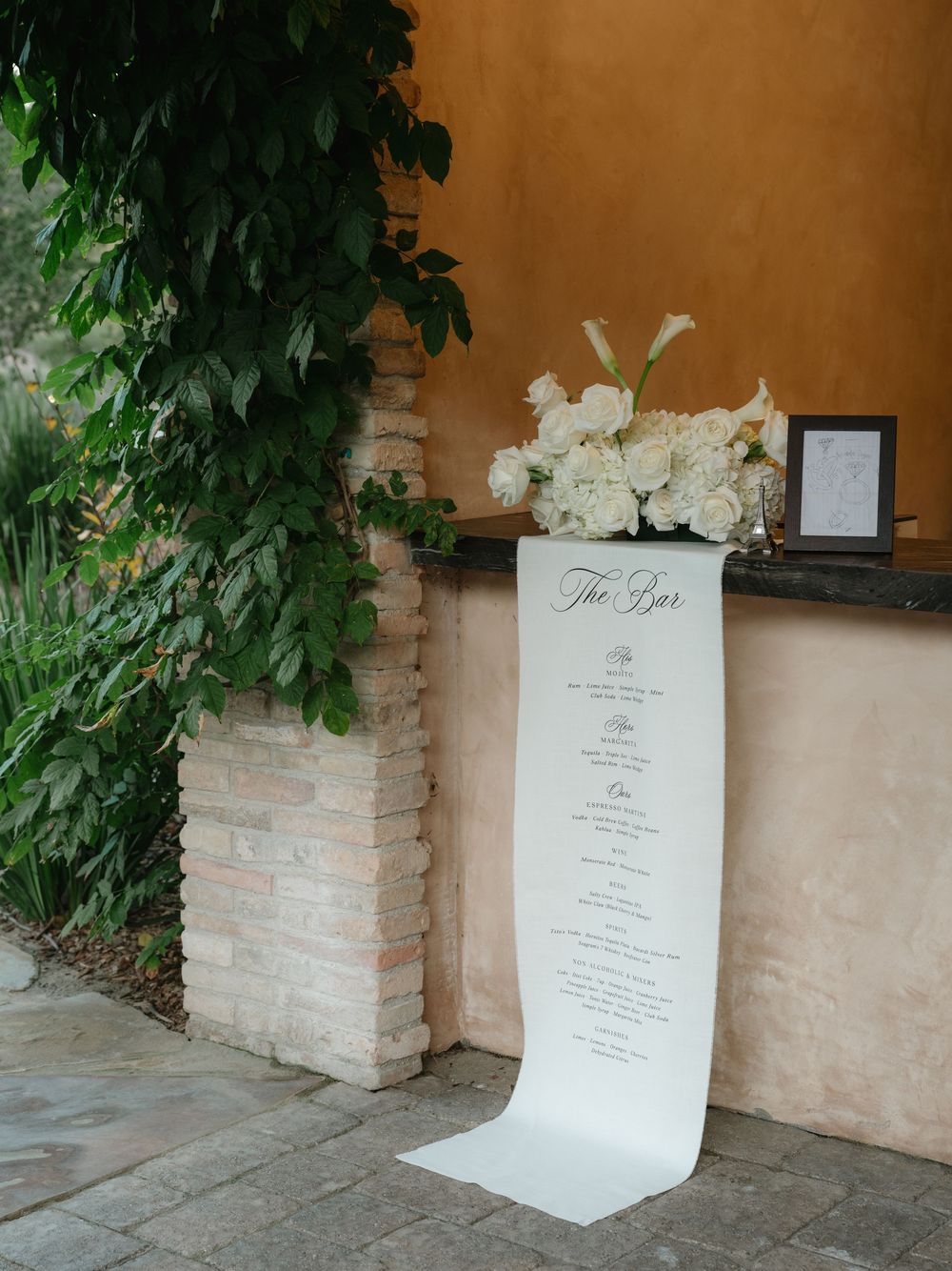 White menu card displayed in an elegant stone planter with white floral arrangements against a terracotta wall.