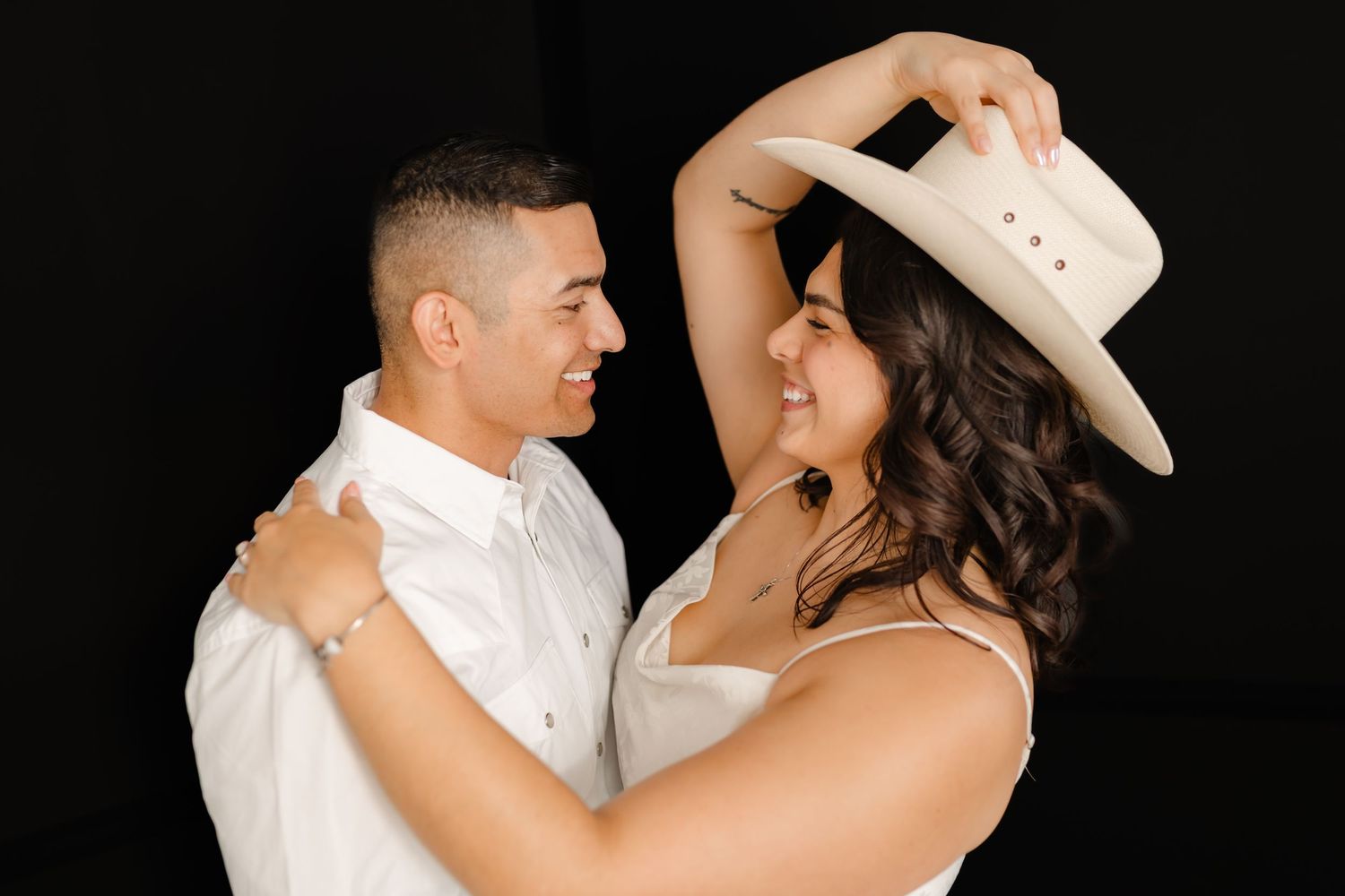 A romantic couple in white attire dance together while the bride wears a cowboy hat during their wedding celebration.