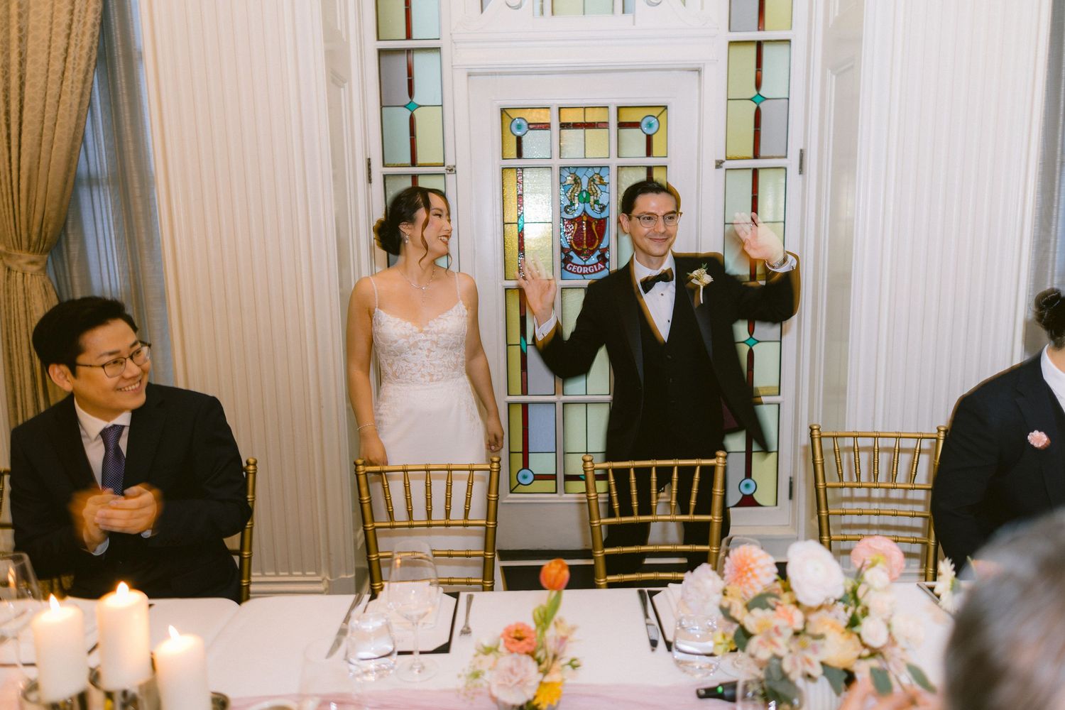 Wedding reception guests seated at elegant tables with pink floral centerpieces and glowing candles in an ornate ballroom.