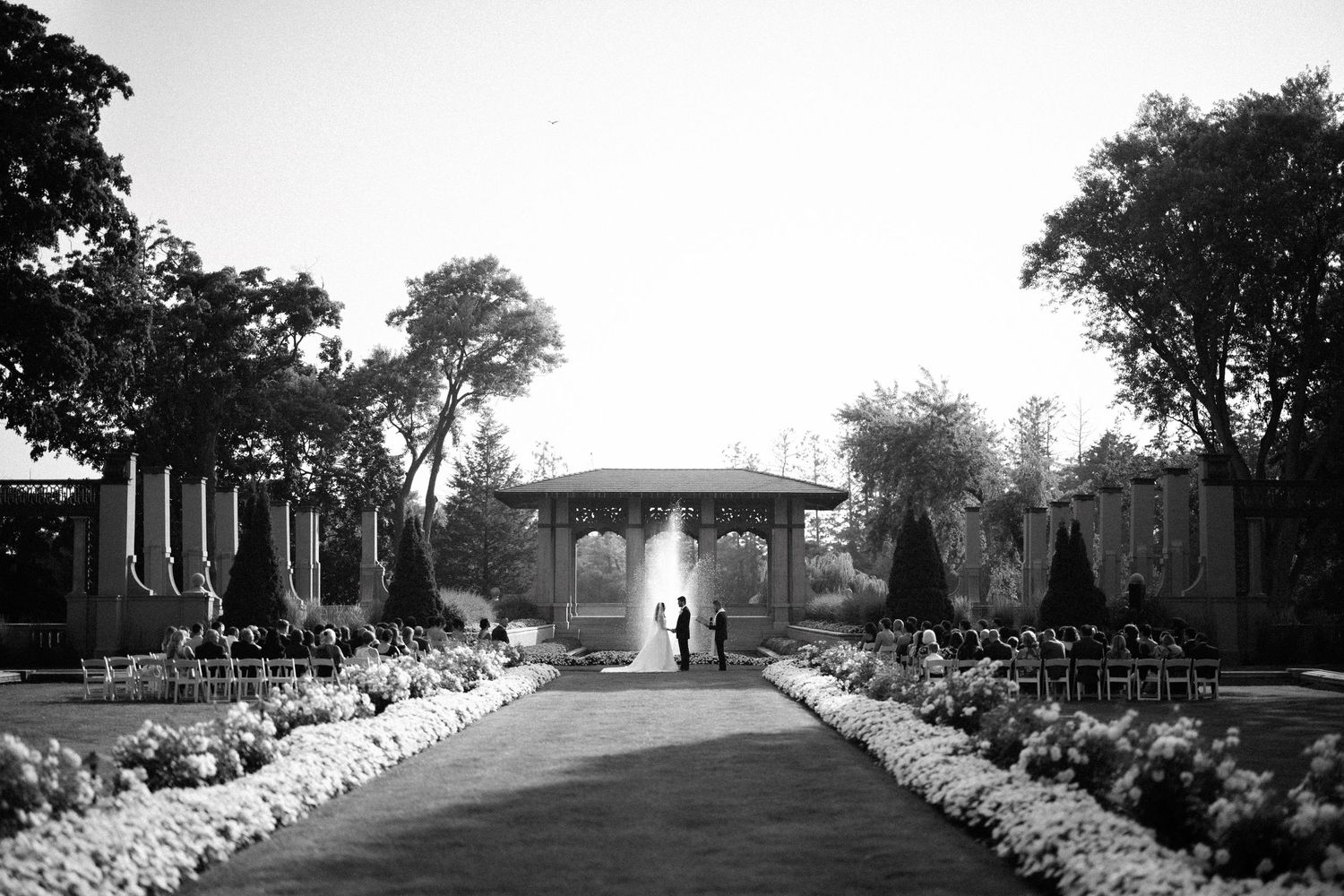 Black and white photograph of a garden path leading to a covered gazebo with flower beds and trees lining the walkway.