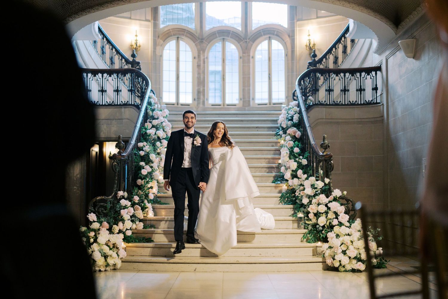 A couple walks down an elegant staircase adorned with white floral arrangements and natural light from tall windows.
