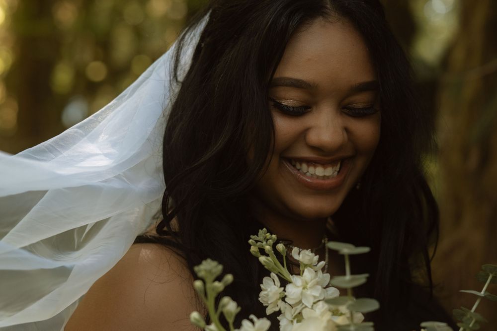 Bride in white veil with flowing dark hair holds white flowers in a lush forest setting.