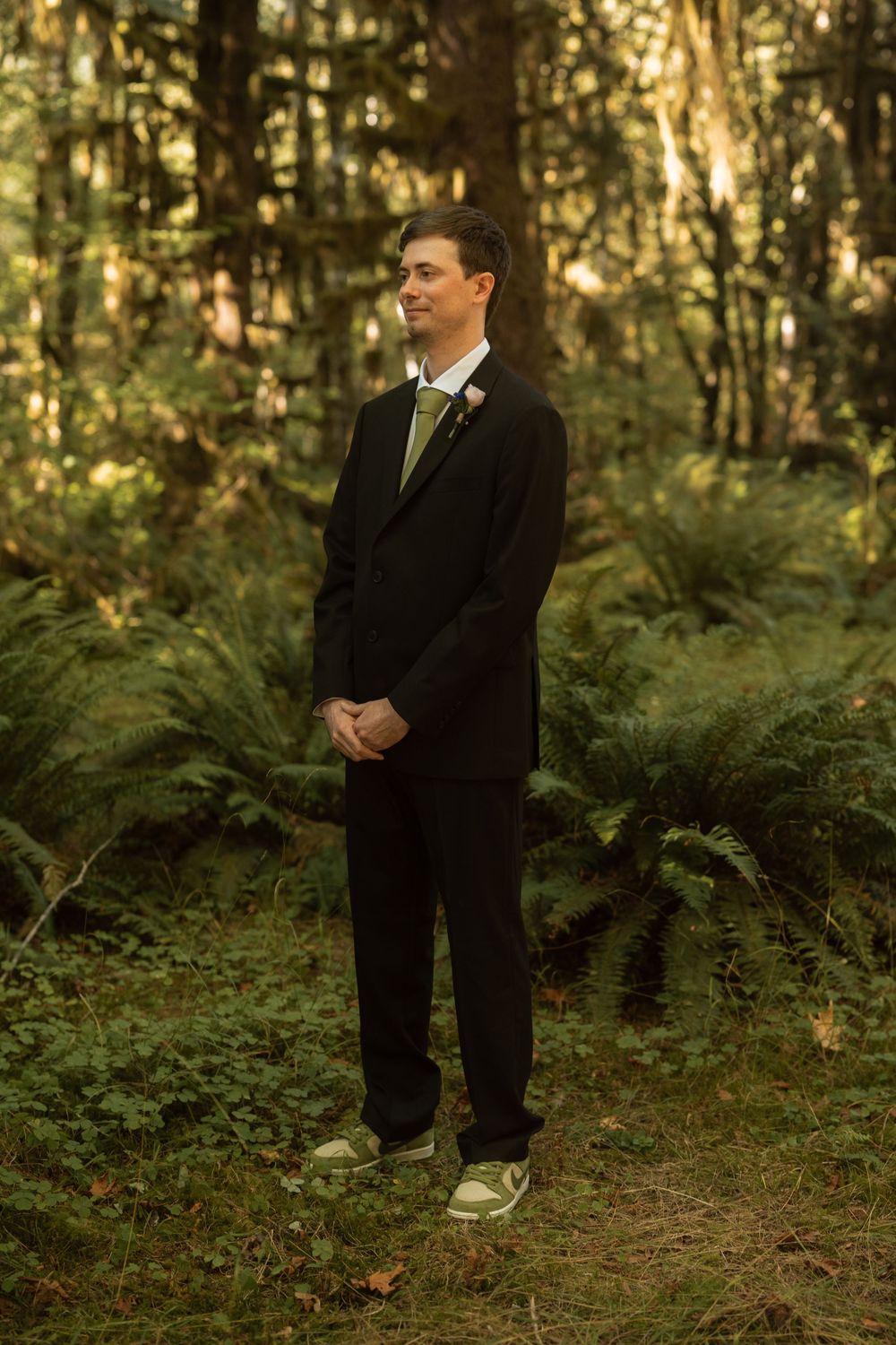 A figure in a black suit and casual shoes stands in a lush forest setting during golden hour.