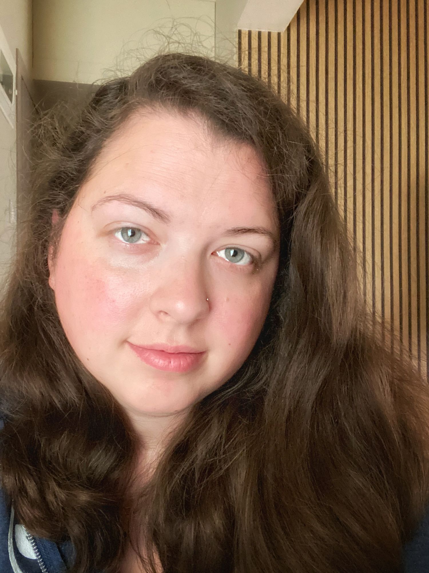 A close-up portrait showing long dark wavy hair and green eyes against a background with wooden slat paneling.