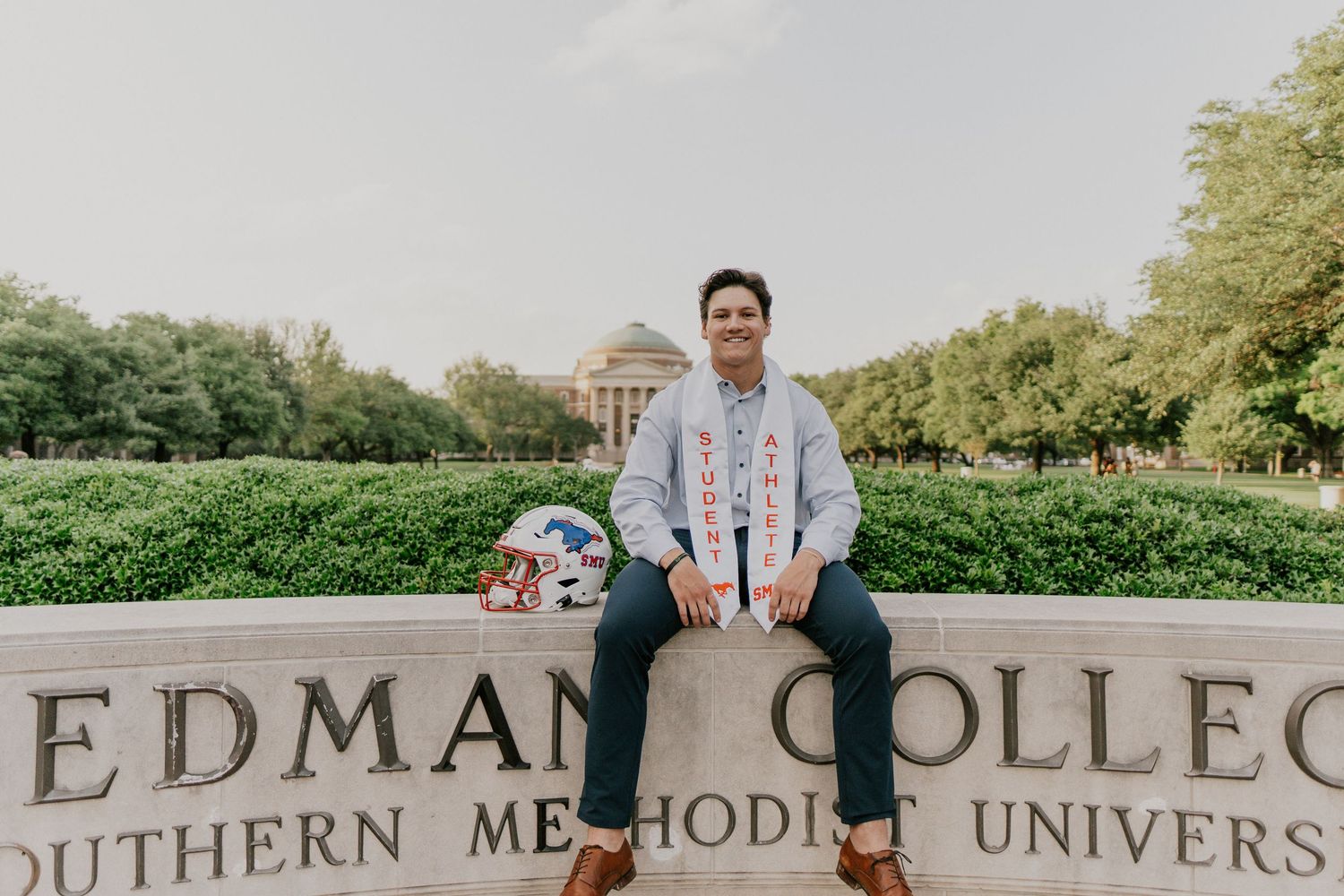 A student sits on a stone sign for Edman College at Southern Methodist University on a sunny day.