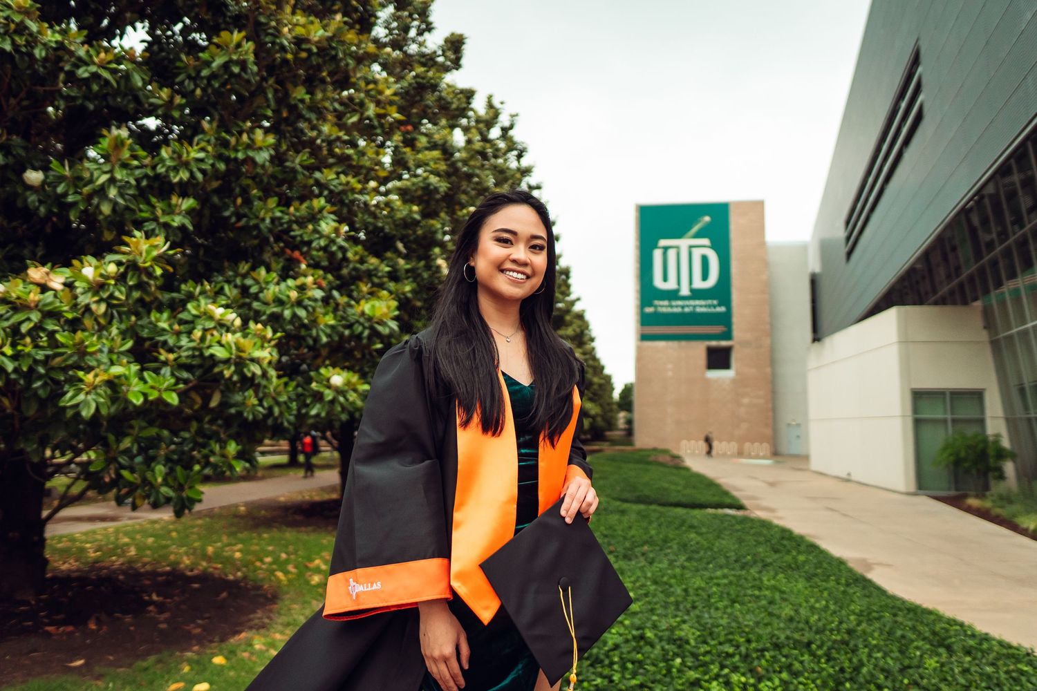 Graduate in orange stole posing near campus building with green landscaping. UTD Graduate Portraits by Eric Aaberg