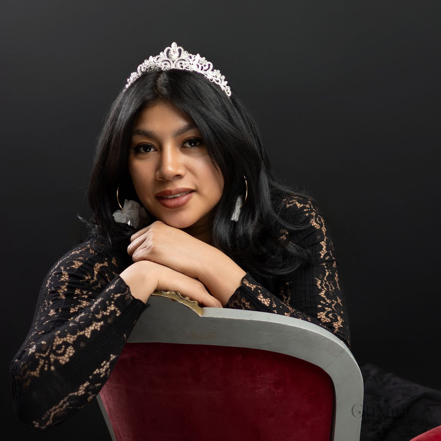 Model in black lace dress poses elegantly with tiara against dark background in glamour photoshoot series.