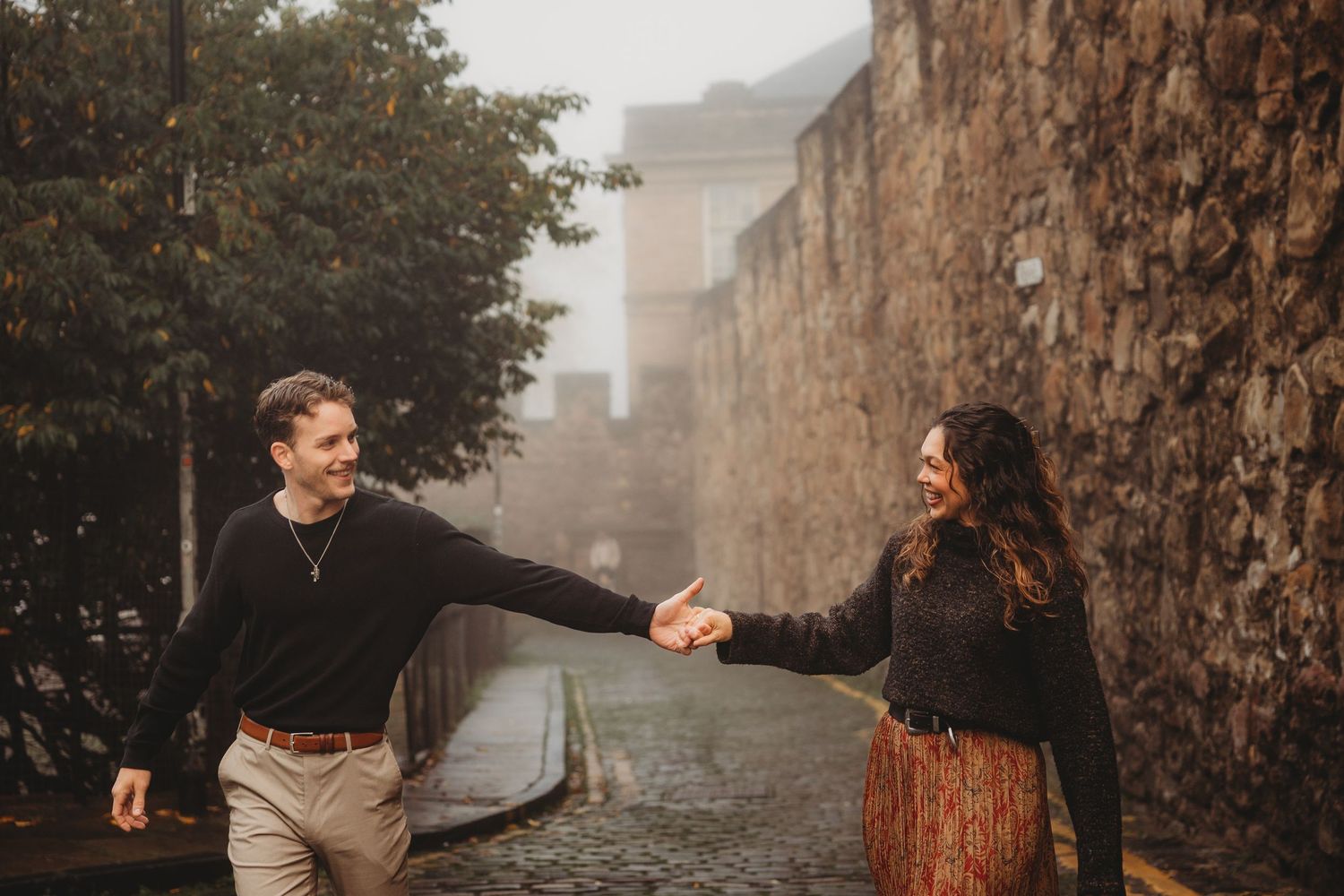 Two people hold hands and laugh while walking along a misty stone wall path in autumn.