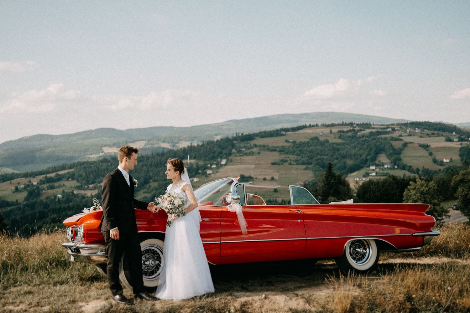 A vintage red convertible car parked on a hilltop with a scenic mountain landscape in the background.