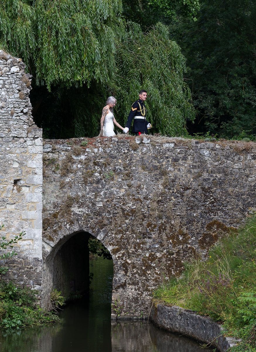 Editorial wedding portrait of bride and groom walking over medieval stone bridge at Allington Castle in Kent