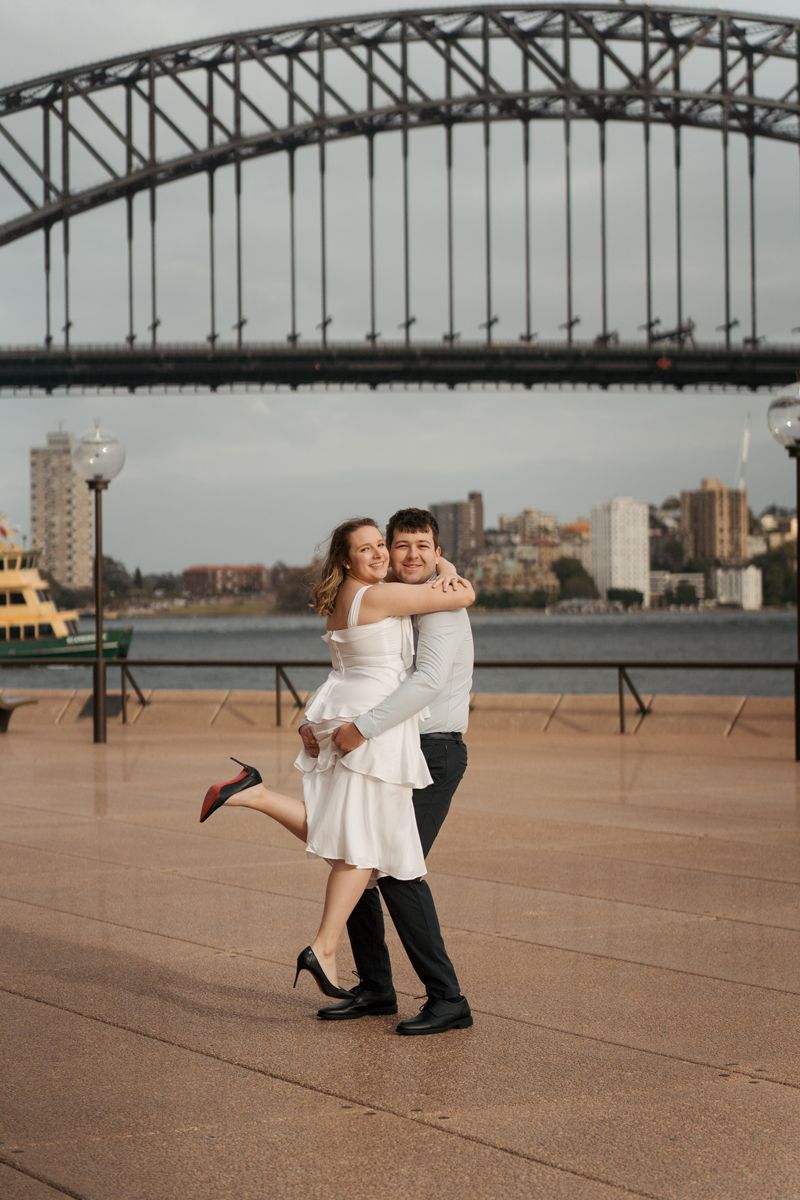 A couple dances and poses together with the Sydney Harbour Bridge silhouetted in the background during their engagement photoshoot in sydney