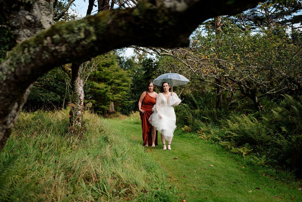 A couple in wedding attire walks through a lush green path surrounded by trees on a sunny day.